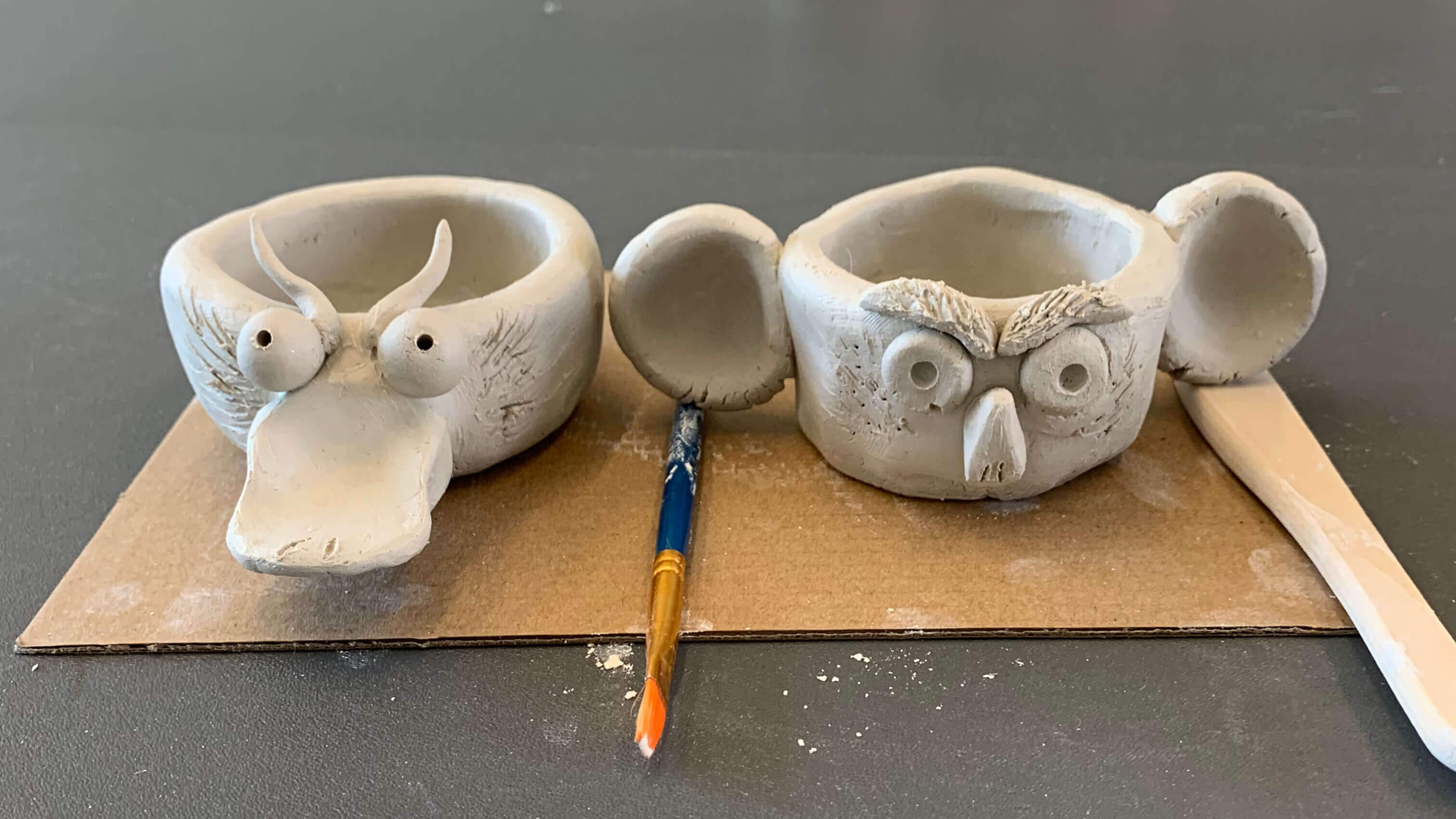 Two animal-shaped bowls on a cardboard surface with a paintbrush and clay tool.