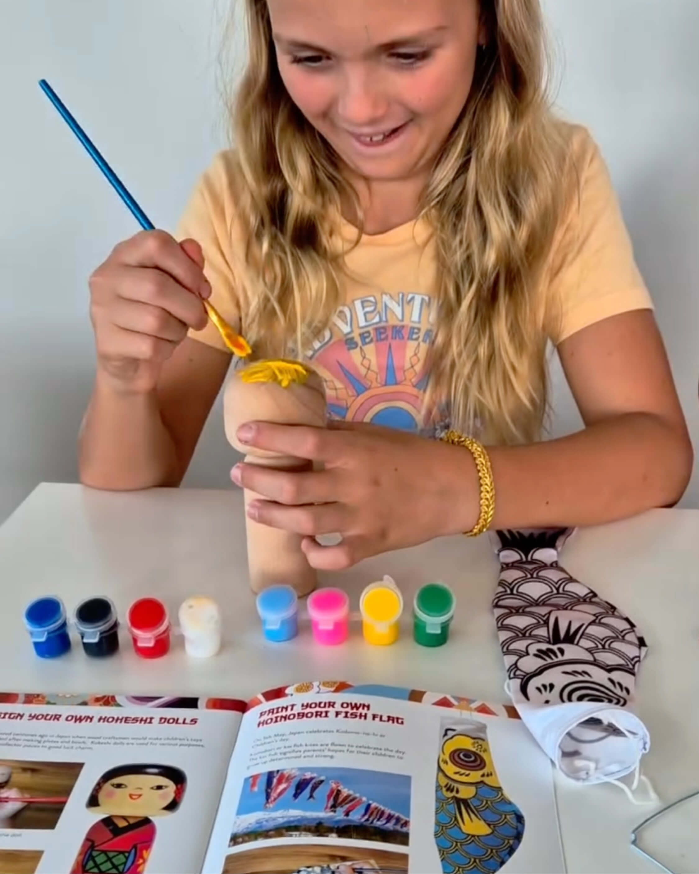 Young girl painting a craft item with colorful paint bottles on a table.